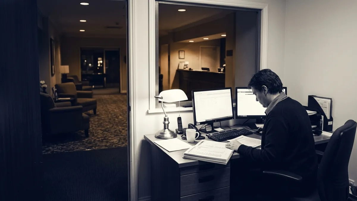 A night auditor working at a back-office desk behind a hotel reception after hours